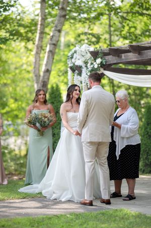 Bride in white strapless gown holds grooms hands at outdoor woodland ceremony with officiant and bridesmaids nearby.