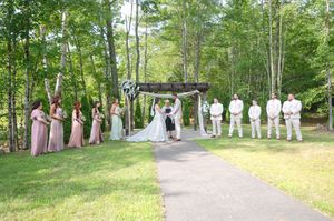 Outdoor wedding ceremony in a forest clearing with bridesmaids in pink and groomsmen in white standing at a rustic wooden arch.