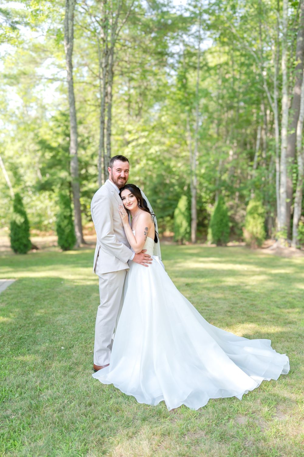 Bride in a flowing white gown with long train embraces groom in tan suit outdoors among tall green trees.