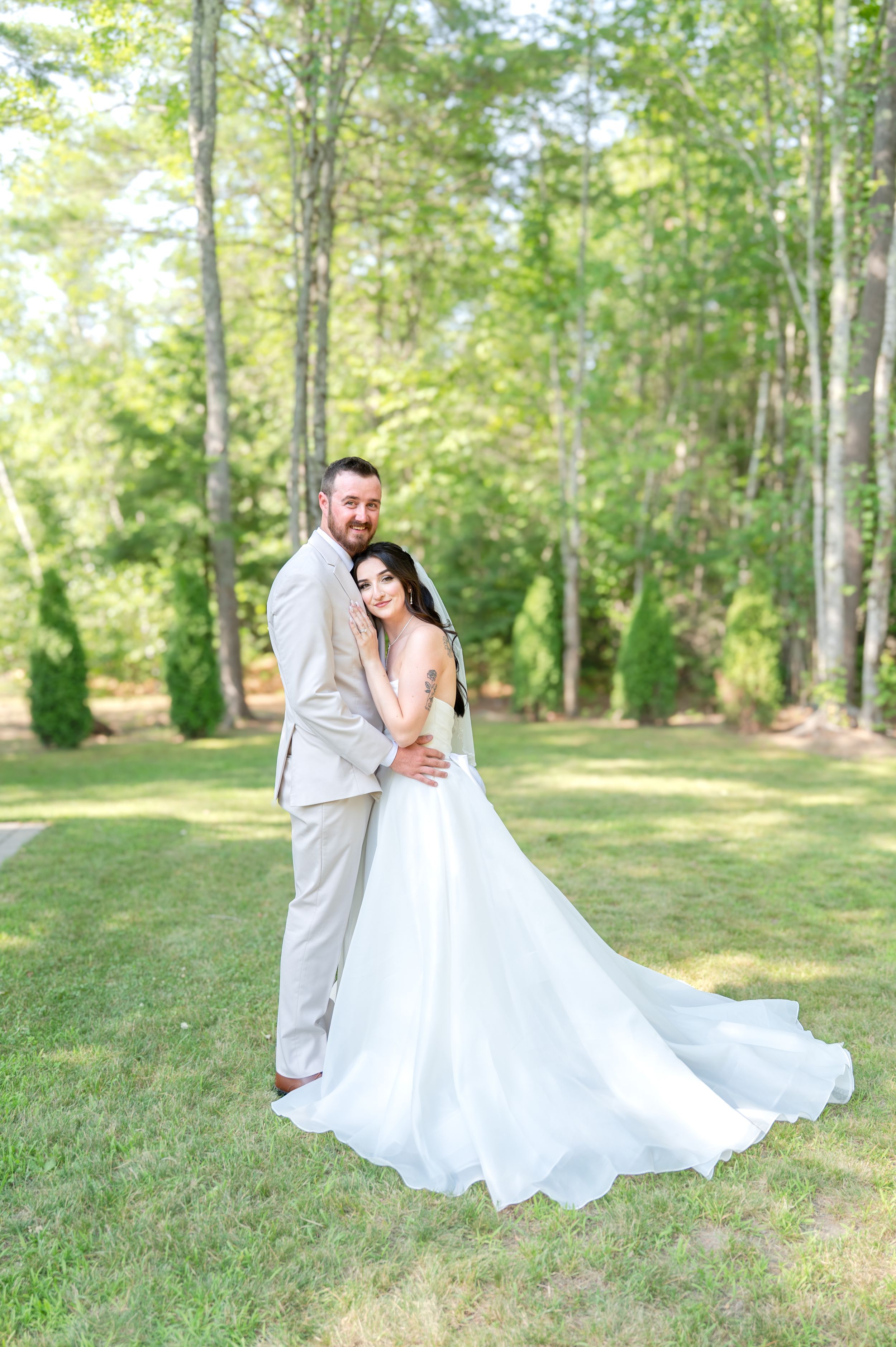 Bride in a flowing white gown with long train embraces groom in tan suit outdoors among tall green trees.