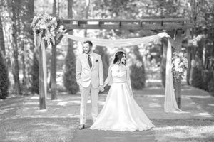 Black and white photo of bride and groom holding hands under a decorated wooden arbor at an outdoor wedding ceremony.