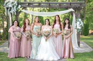 Bride with four bridesmaids in mismatched pink and sage gowns holding bouquets in front of a rustic wooden wedding arch.