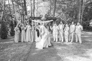 Black and white photo of bride and groom dipping during kiss while wedding party cheers beside a wooden arbor.