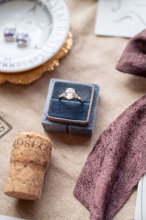 Diamond engagement ring in a blue velvet box beside a wine cork and mauve ribbon on linen fabric.