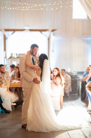 Bride and groom share their first dance in a warmly lit barn reception venue surrounded by seated wedding guests.