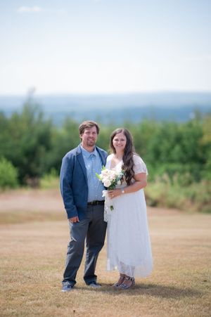 Bride in white dress holding bouquet stands with groom in blue blazer on grassy hilltop with scenic landscape behind.