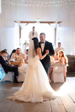 Bride in a backless gown dances with a man in a black tuxedo in a white barn reception hall.