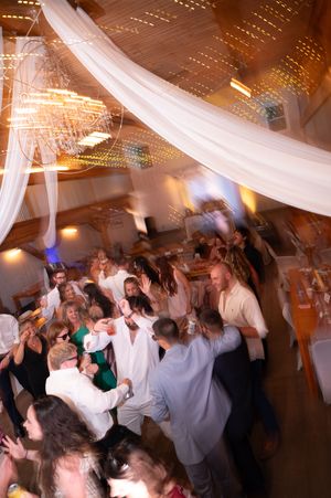 Energetic wedding reception dance floor packed with guests celebrating under white draped fabric and sparkling string lights.