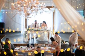 Bride and groom sit at a sweetheart table in an elegantly decorated barn reception hall with bokeh string lights.