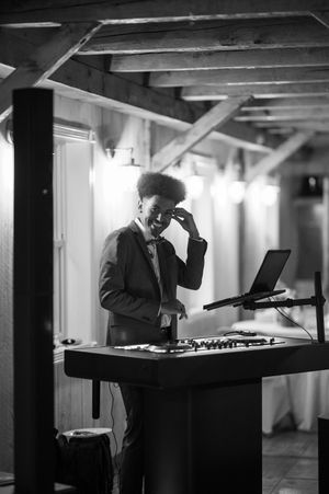 DJ smiling at his equipment setup in a rustic venue, captured in black and white photography.
