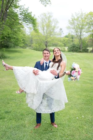 Groom carrying bride in white lace wedding dress and veil while she holds a pink bouquet on a green lawn.