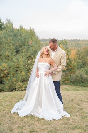 Groom in tan suit embracing bride in strapless white gown with long veil among autumn trees outdoors.
