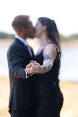 Couple in black wedding attire share a kiss while holding hands near a sunlit waterfront setting.
