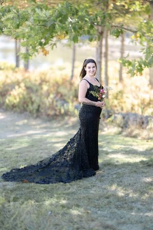 Bride in stunning black lace mermaid gown with train holds a colorful bouquet among autumn trees.