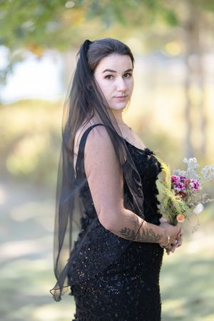 Bride in black sequin gown with black veil holds a wildflower bouquet in a sunlit outdoor setting.