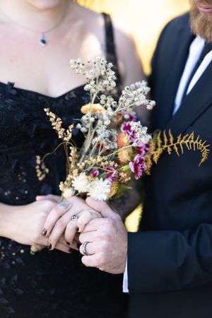 Bride and groom hold a wildflower bouquet together, showcasing their wedding rings in close-up detail.