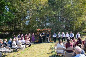 Outdoor wedding ceremony in a forest clearing with wedding party at floral arch and guests seated in white chairs.