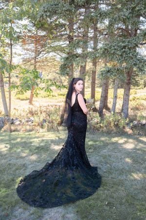 Woman in elegant black lace mermaid gown with long train poses among trees in a sunlit outdoor setting.