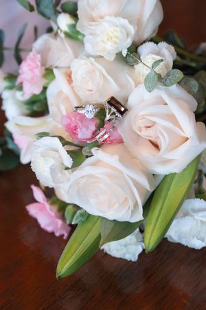 Wedding rings rest among blush roses, pink carnations, and white flowers in a bridal bouquet arrangement.
