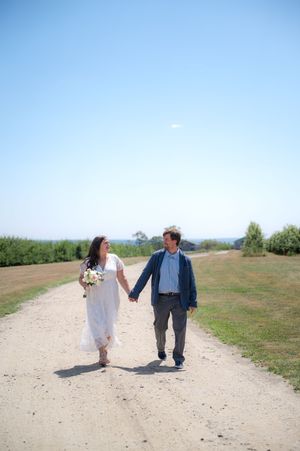 Bride and groom walk hand in hand down a sunny gravel path surrounded by greenery on their wedding day.