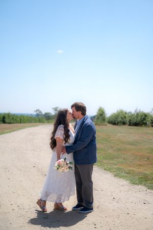 Bride and groom share a romantic kiss on a sunlit gravel path with blue sky and green fields behind them.