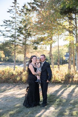 Family of three poses outdoors in formal attire, woman in black sequin gown holding toddler beside suited man.