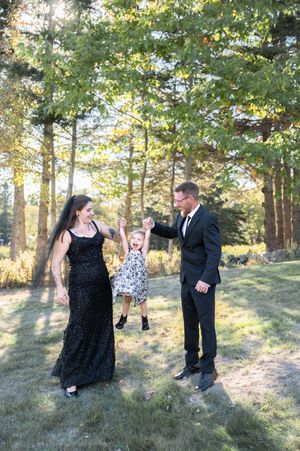 Couple in formal wear joyfully swings young child between them in a sunny tree-lined outdoor setting.
