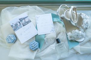 Elegant wedding flat lay featuring invitations, blue ring box, perfume, heels, and floral accessories.