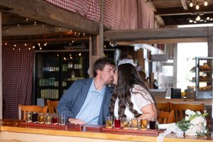 Newlywed couple shares a kiss at a rustic wooden bar surrounded by warm string lights and wine bottles.