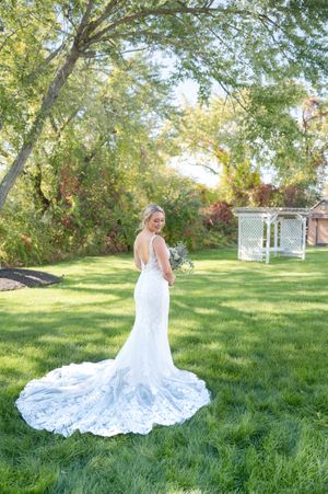 Bride in backless lace mermaid gown with long train stands in green garden near white gazebo.