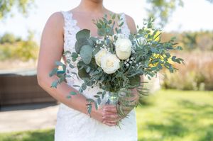 Bride holds lush bouquet of white roses and eucalyptus greenery in lace wedding gown outdoors.