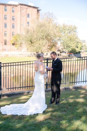 Groom reads vows to bride near iron fence by river with historic brick building in background.