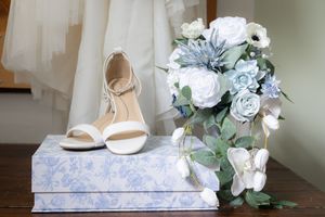 Wedding flat lay featuring white heels on a floral box beside a cascading blue and white bridal bouquet.