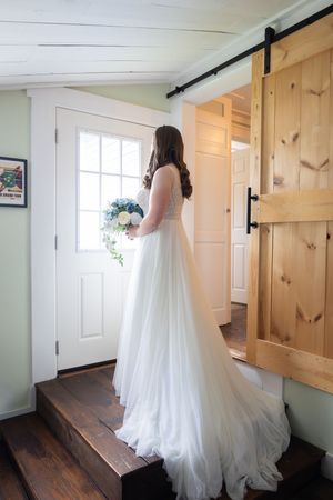 Bride in white gown holding blue and white bouquet standing by a door near a rustic barn sliding door.