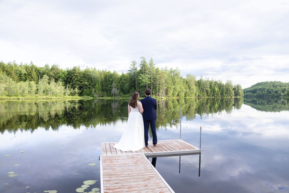 Bride and groom standing at the end of a wooden dock overlooking a perfectly still reflective forest lake.