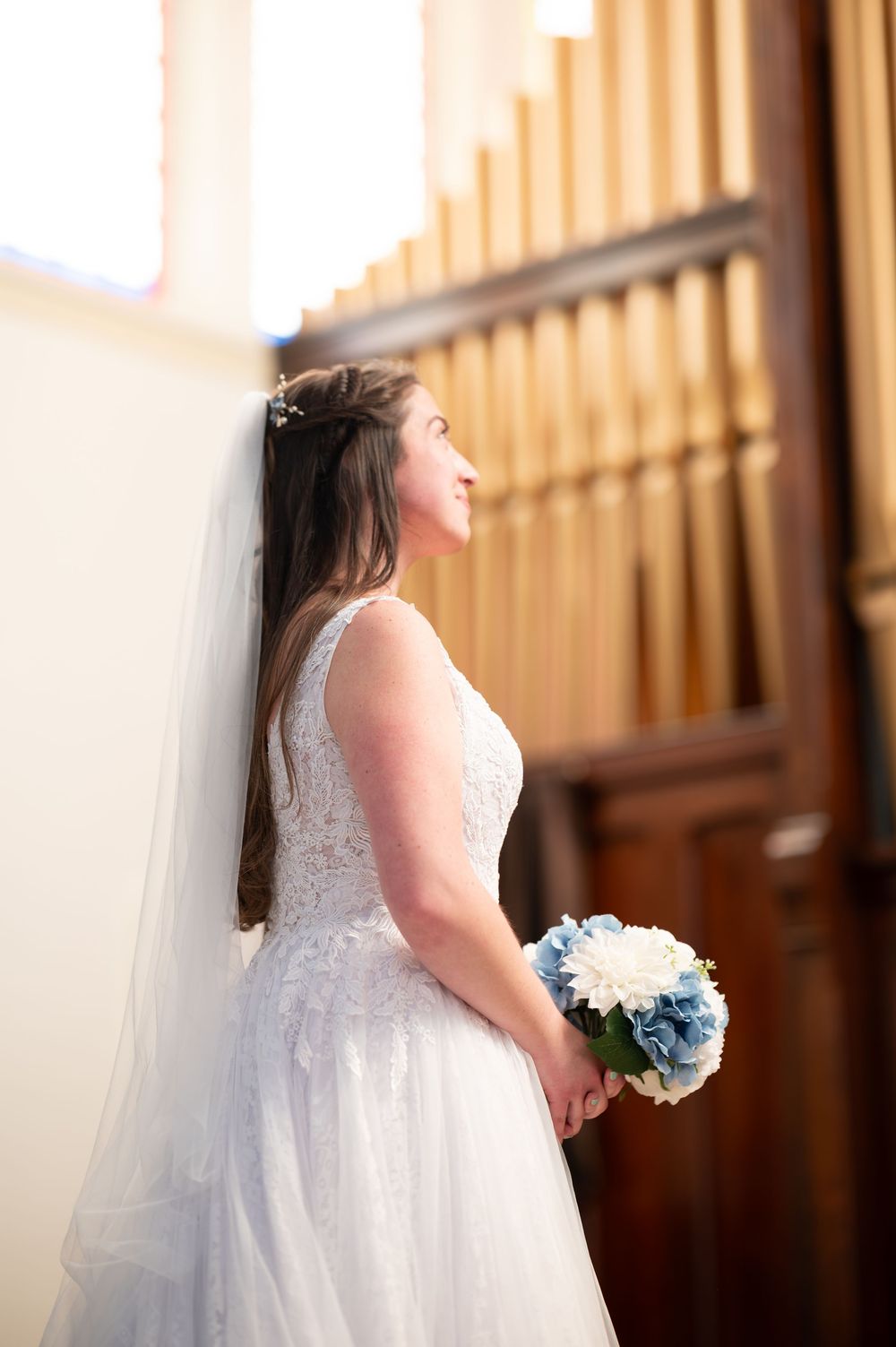 Bride in white lace gown holding blue and white bouquet, leaning against wall near church organ pipes.