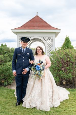 Bride in lace ballgown and groom in military dress uniform pose together in front of a white gazebo.