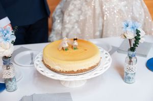 Cheesecake wedding cake on a white pedestal stand adorned with miniature bride and groom cake toppers.