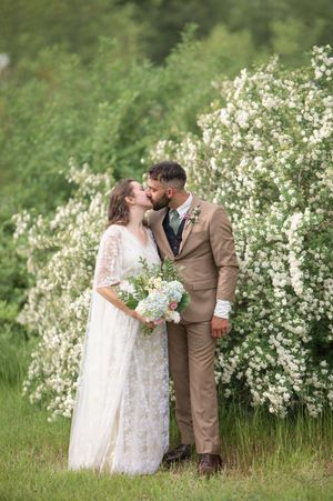 Bride and groom share romantic kiss in front of blooming white flower bushes outdoors.