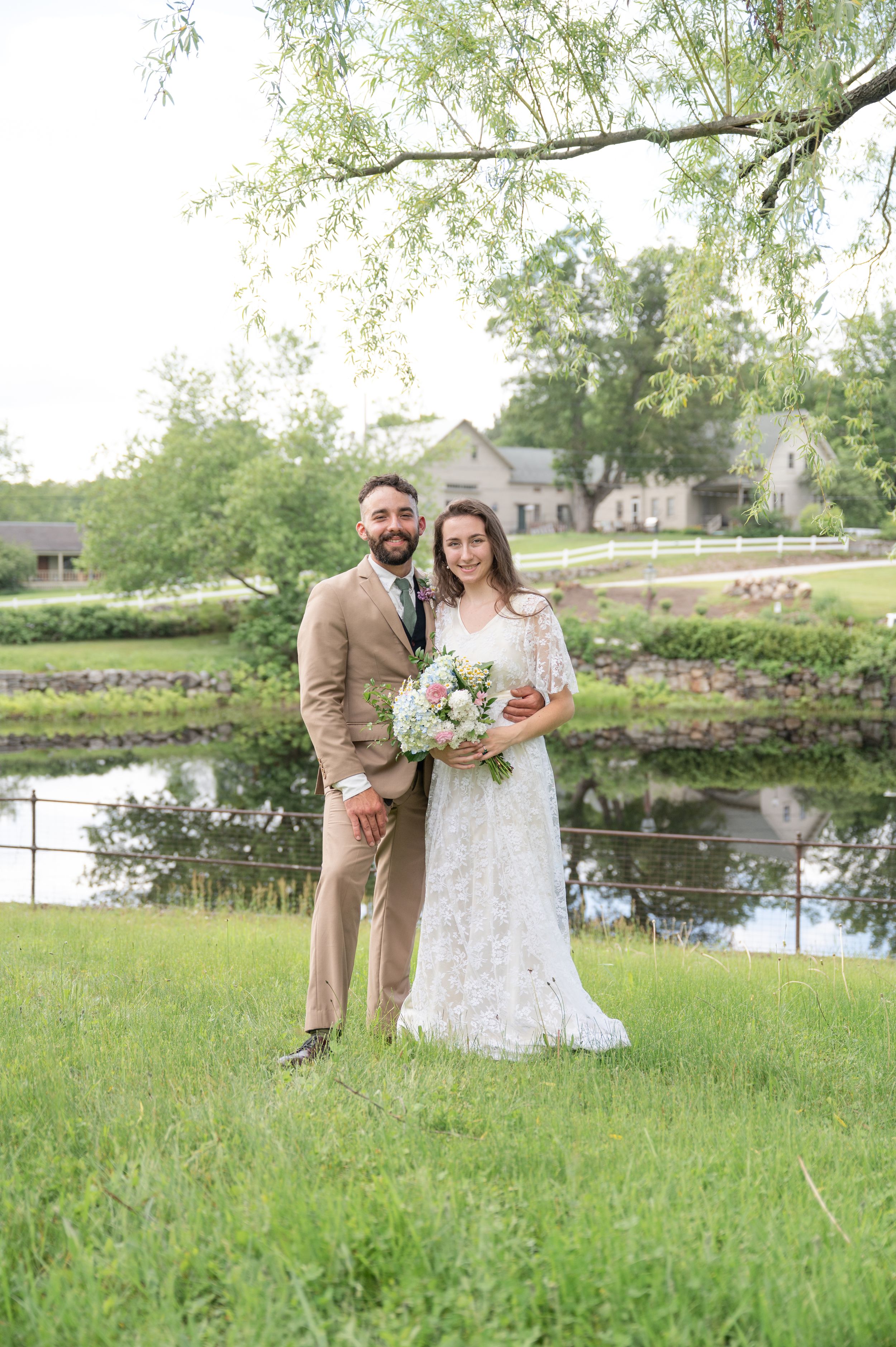 Bride and groom portrait standing on green grass near reflective pond under willow tree.