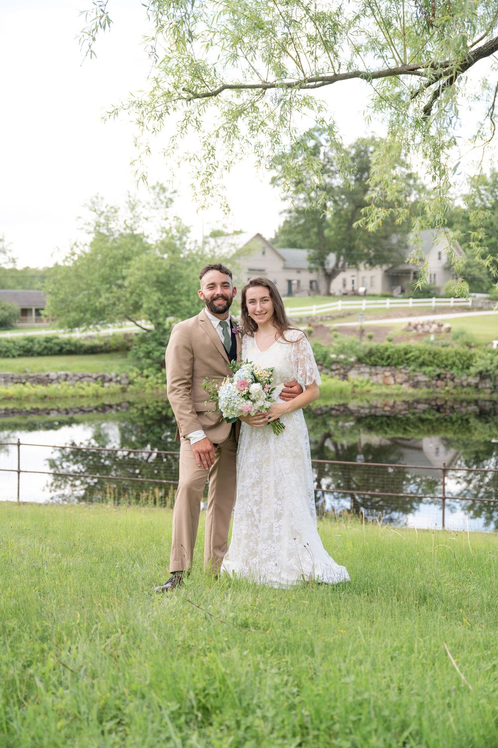Bride and groom portrait standing on green grass near reflective pond under willow tree.