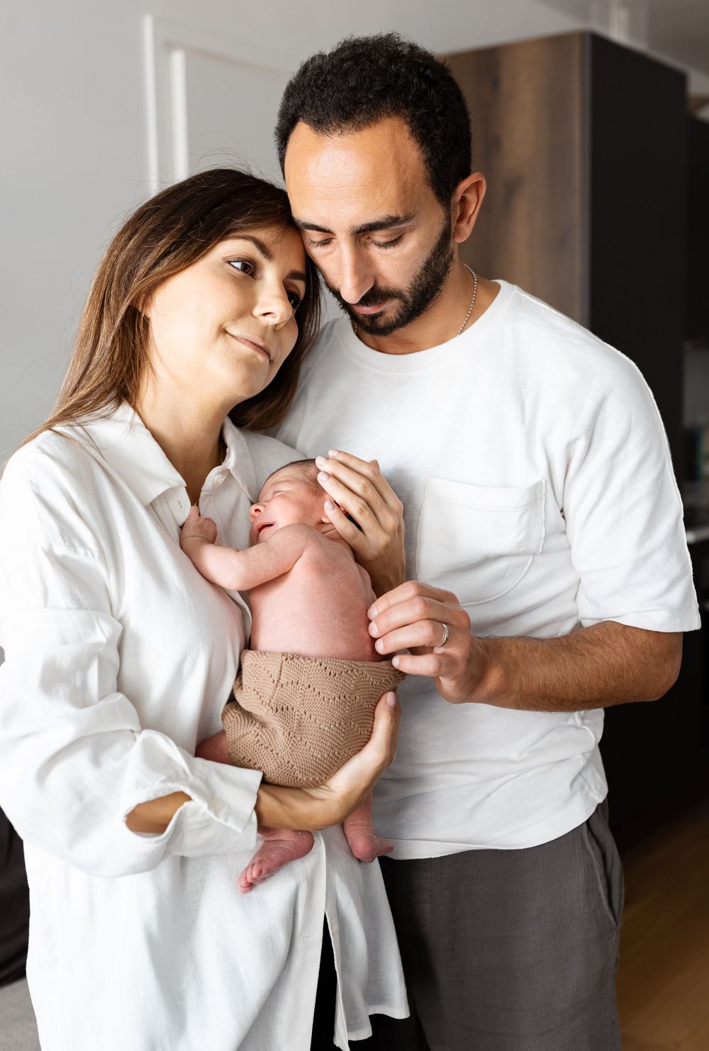 An intimate portrait of new parents sharing a tender moment with their newborn in matching white attire.