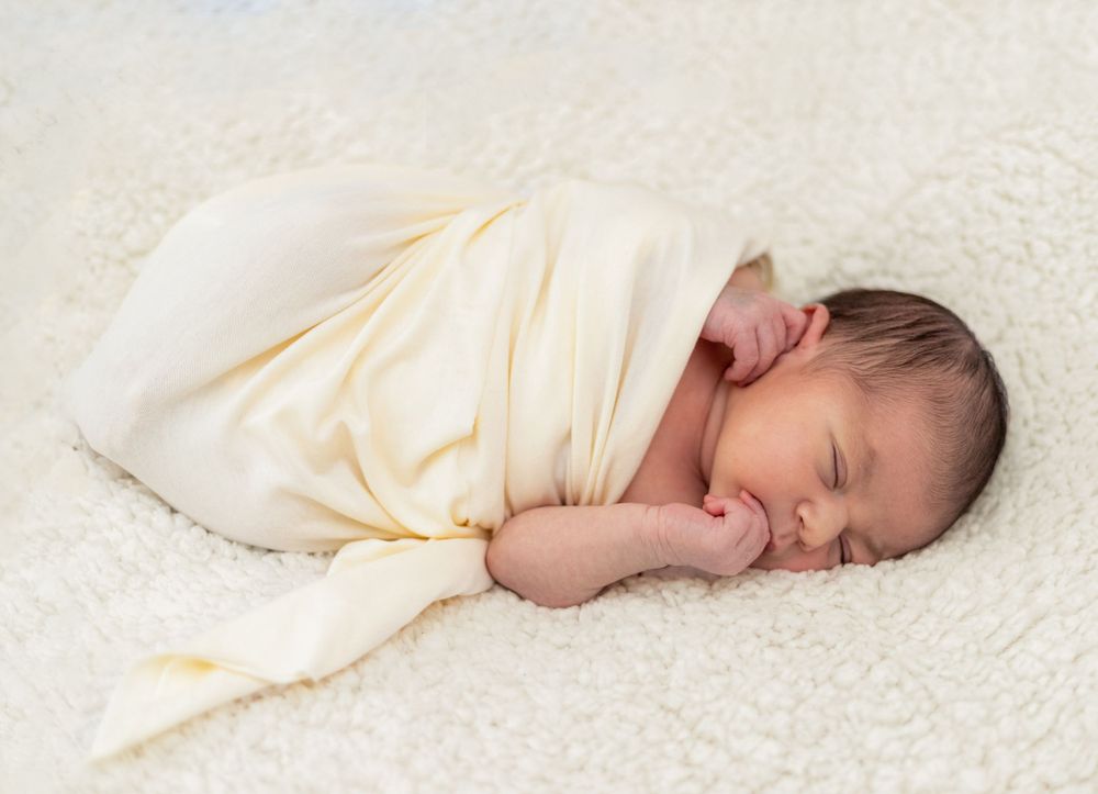Peaceful newborn baby sleeping swaddled in cream colored blanket on textured white surface.