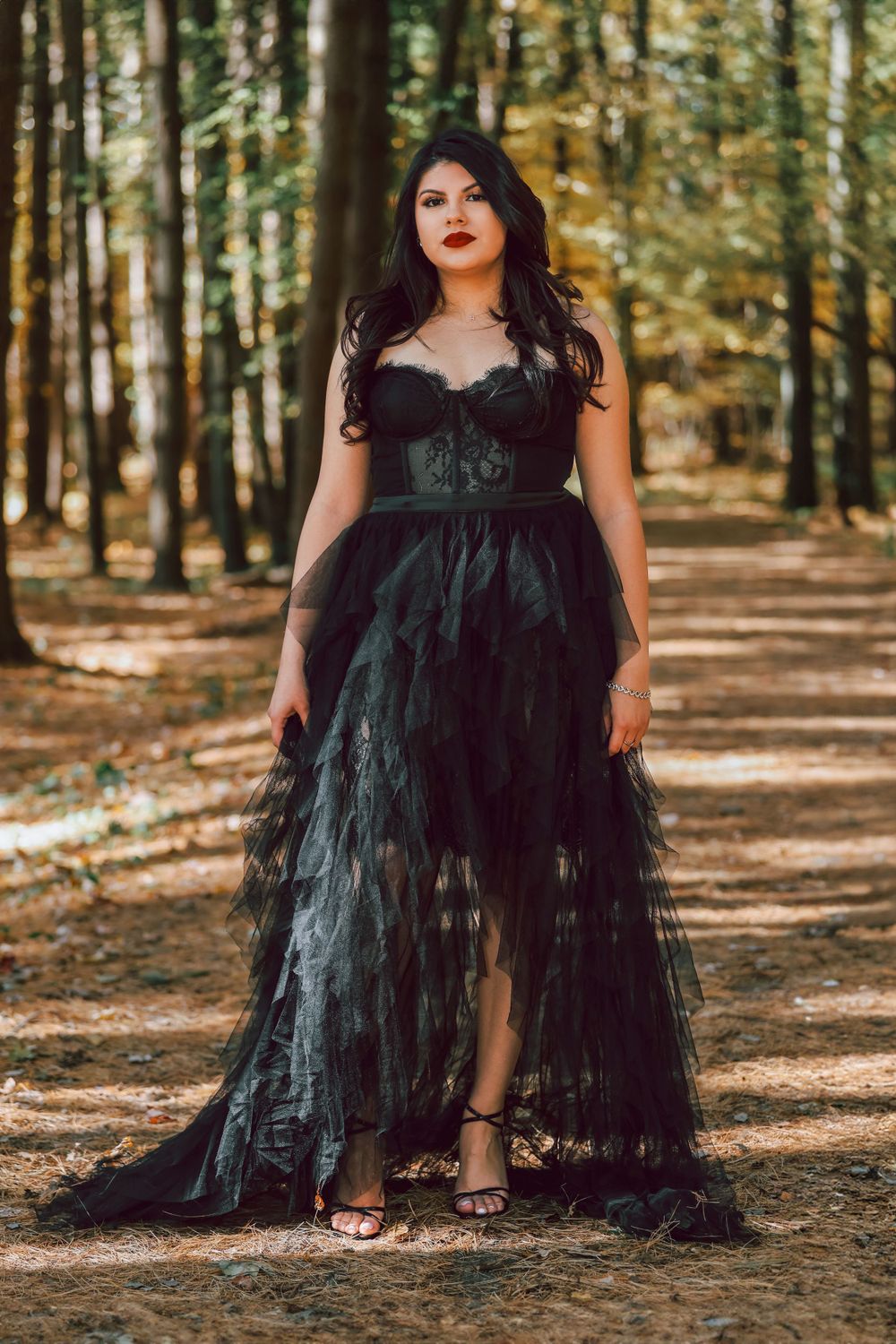 A woman in a dramatic black tulle gown poses in a sunlit forest setting.