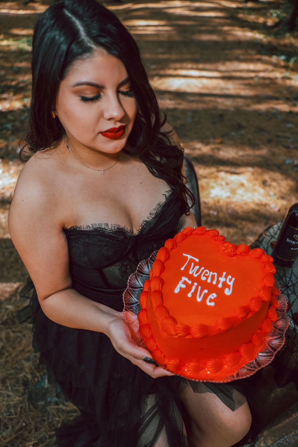 A woman in a black dress holds a red cake with "Twenty Five" written on it in a forest setting.