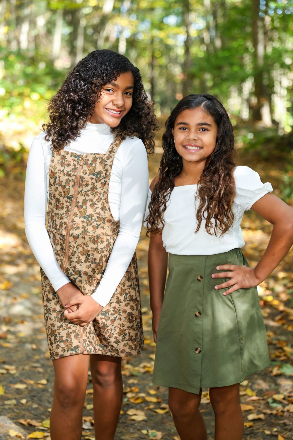 Two young girls in fall outfits smile together on a wooded path.