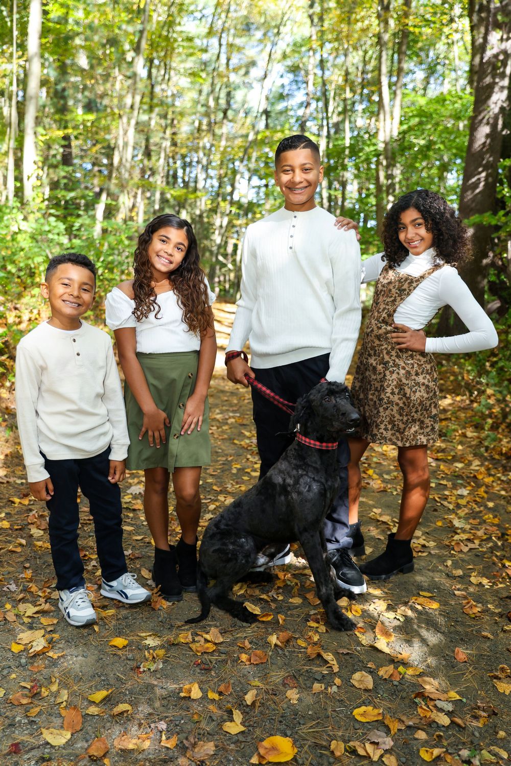A group poses with their black dog on a leaf-covered forest path during autumn.