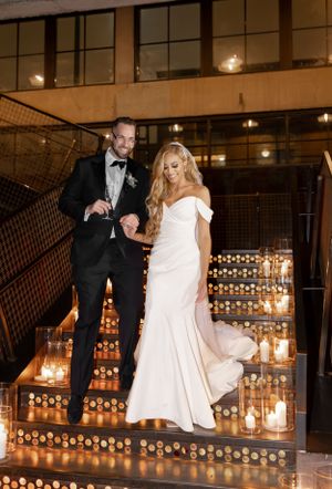 Newlyweds descend candlelit stairs in formal attire at an industrial-chic wedding venue.