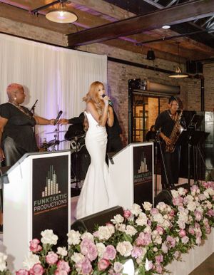 Bride in white gown performs with live band on stage surrounded by floral decorations.