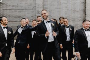 Groomsmen in black tuxedos laughing together against brick wall background.
